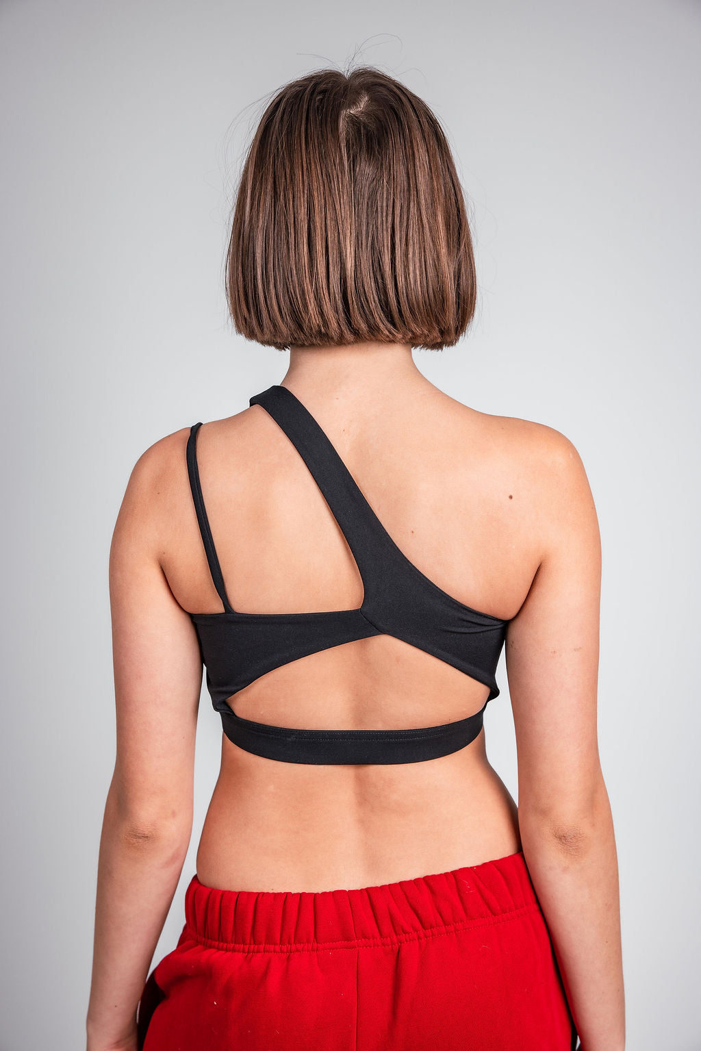 Woman wearing black CAN. Syndicate Asymmetrical Cut-Out Crop Top and red track pants facing grey back drop 