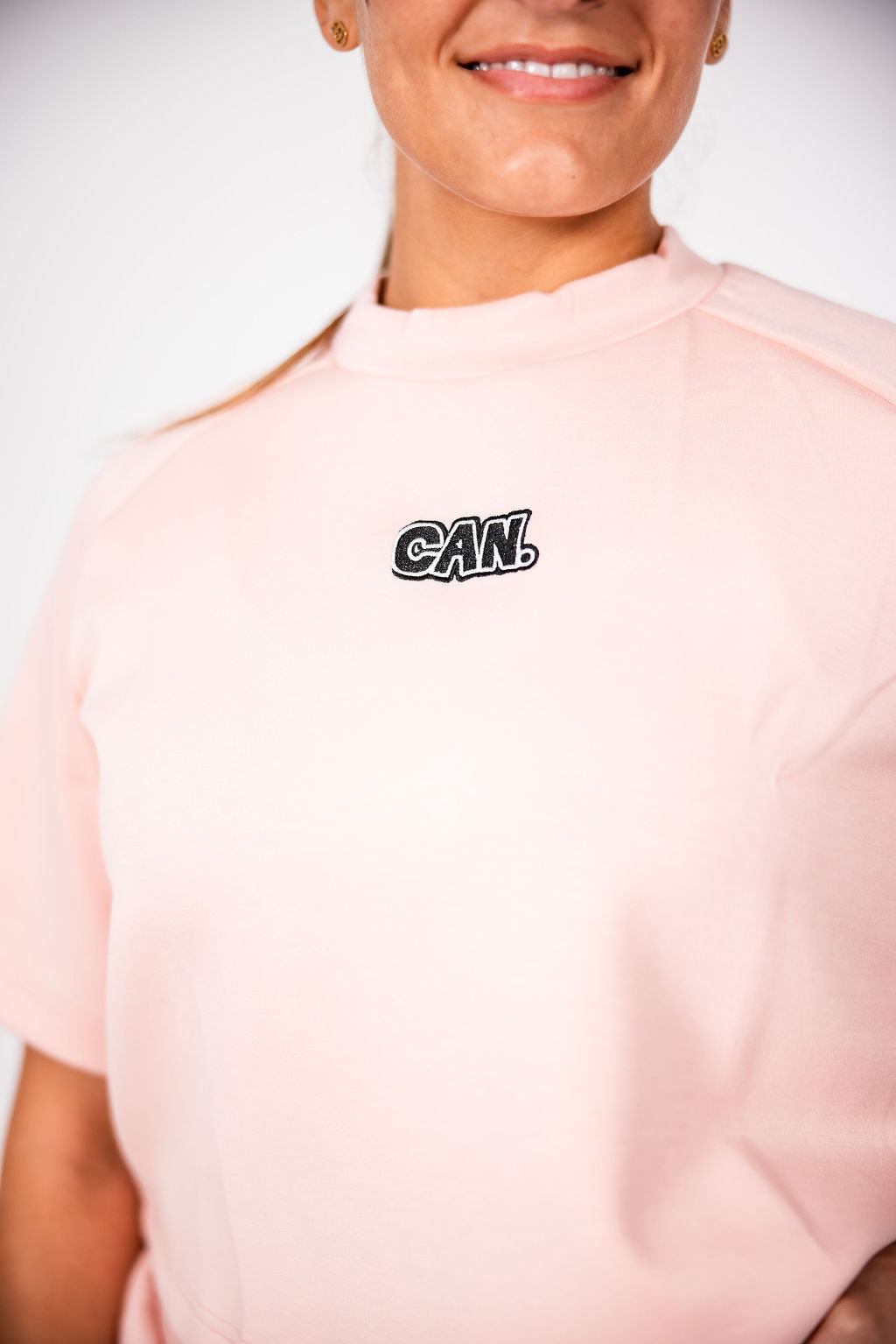 A close-up of a person wearing a light pink Can. Syndicate Comfort  Cropped Tee against a light grey background