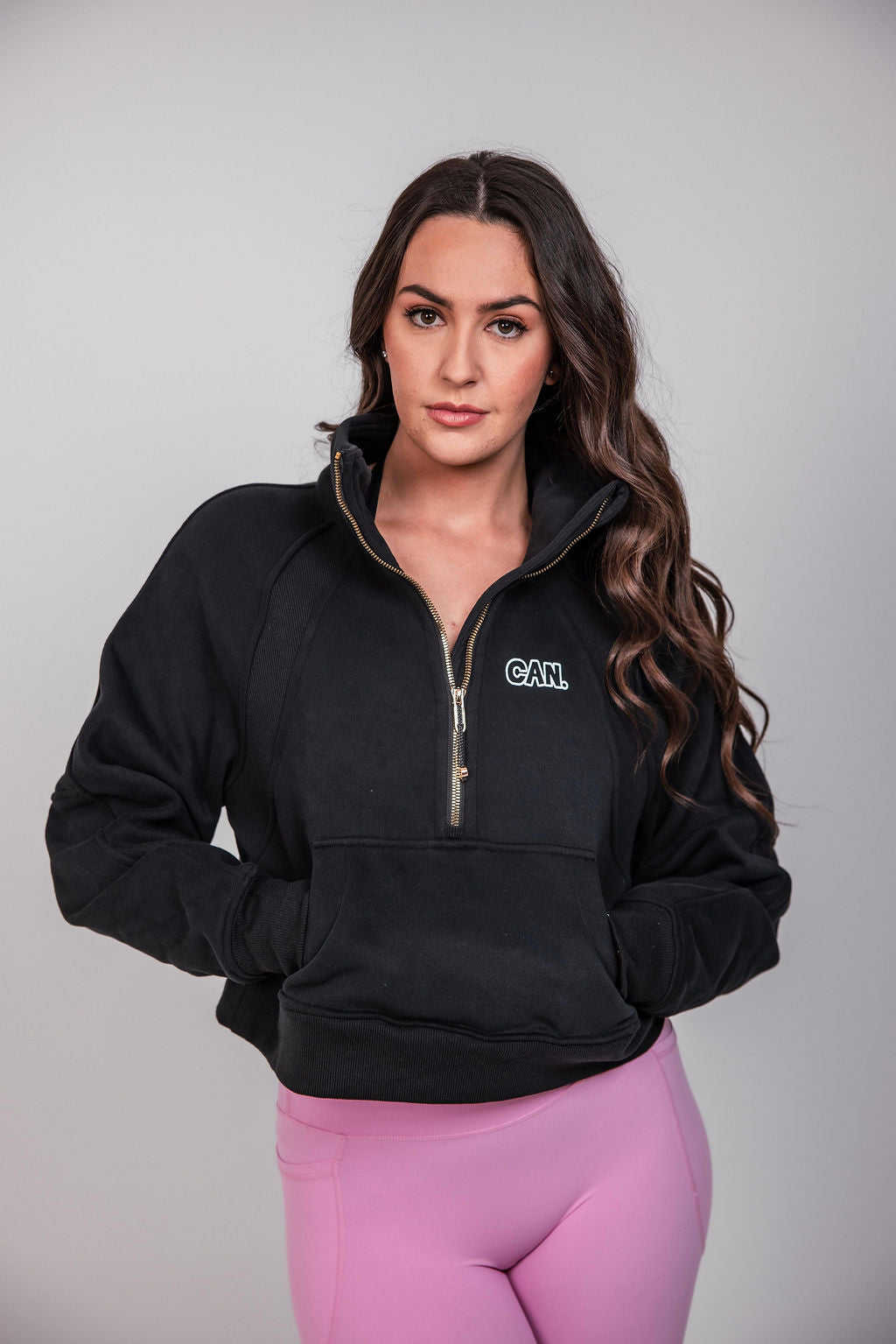 A woman modelling a Can. Syndicate Cropped Zip Jumper  with a white embroidered 'CAN.' logo on the left chest, paired with pink leggings, against a solid light grey background.