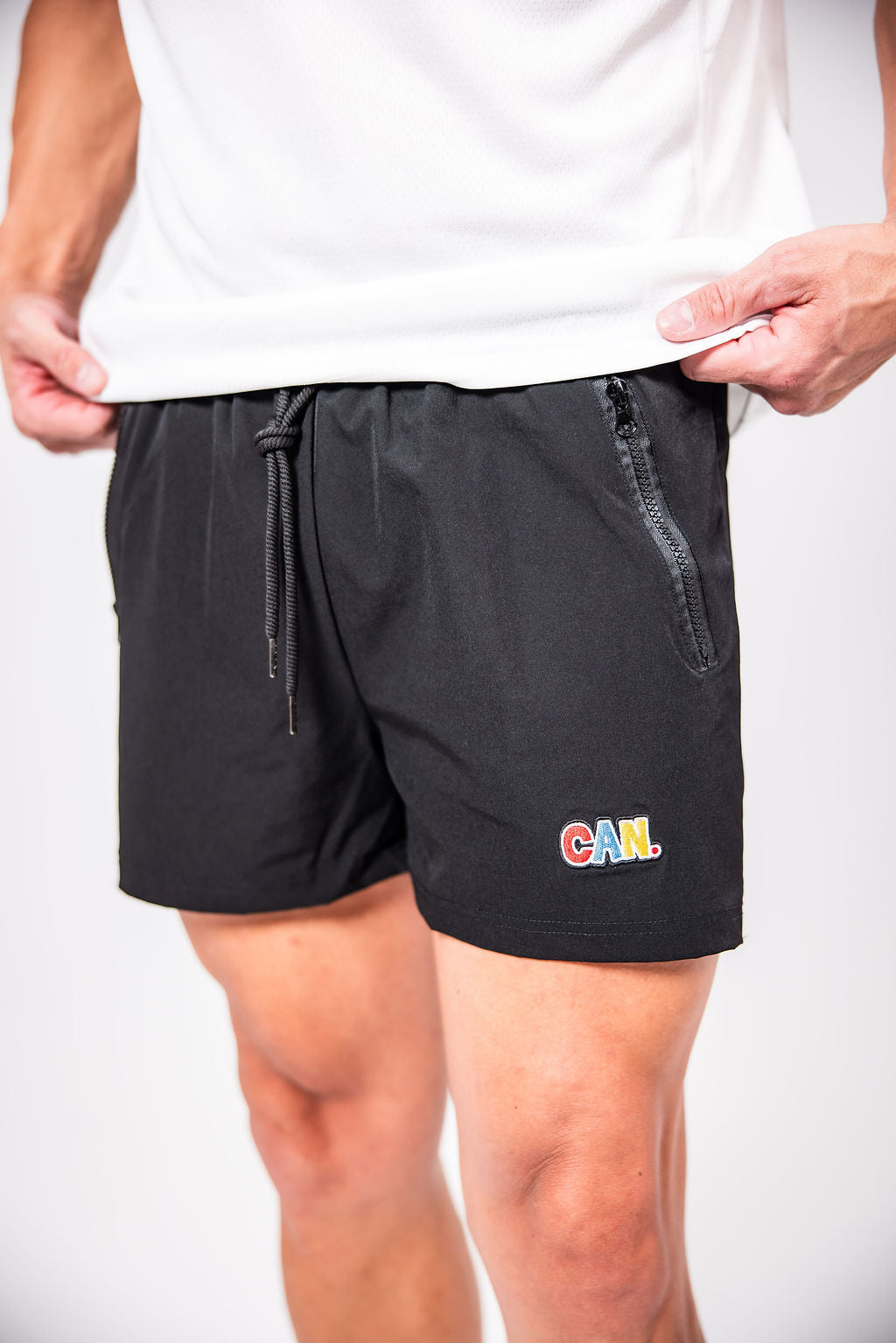 A man wearing a white shirt and black CAN. Men's gym shorts with a red, blue and yellow logo that says CAN. on a white background