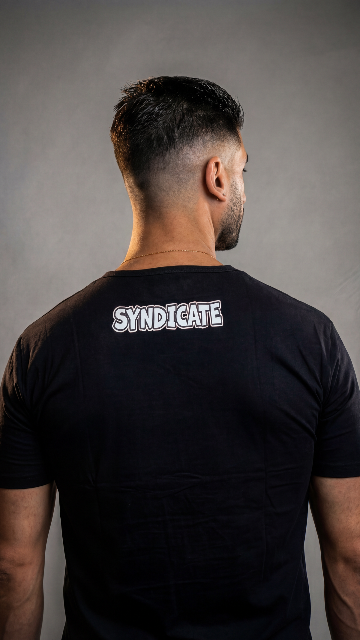 Man wearing a black CAN. Syndicate Men's Fitted Tee with 'SYNDICATE' logo on the back against a grey background
