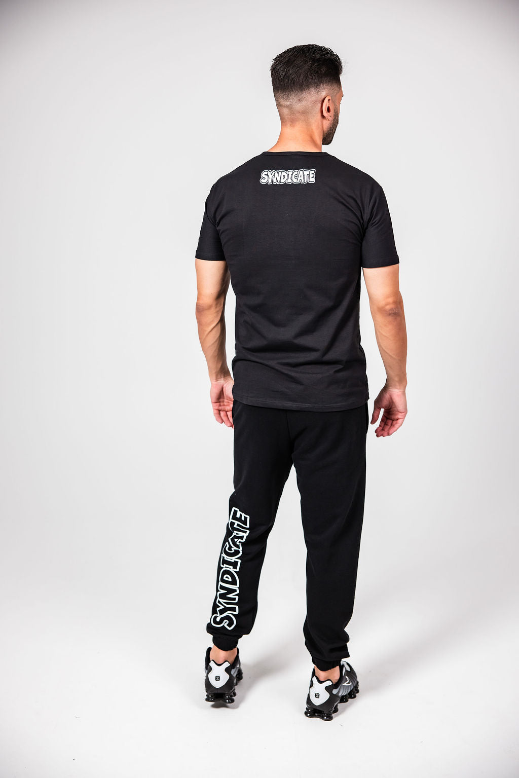 James Webb wearing a black CAN. Syndicate Men's Fitted Tee  and black statement cuffed track pants with 'SYNDICATE' branding on a white background