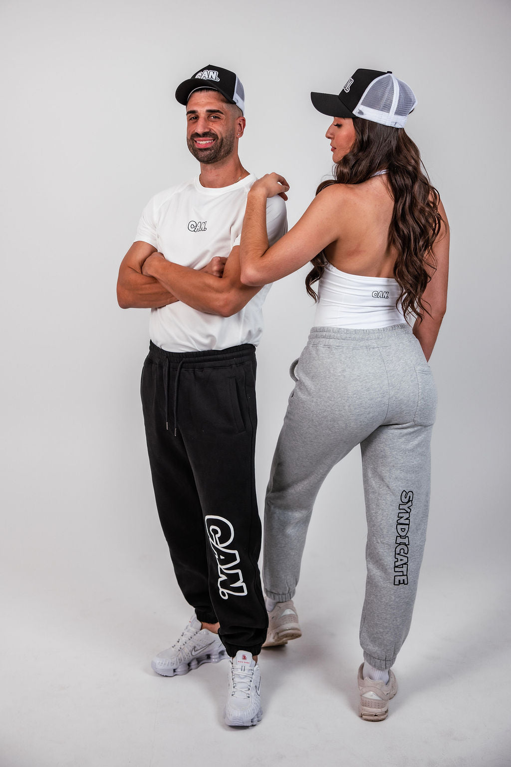 A man and a woman modelling matching white athletic apparel with black and gray accents. The man wears a white short-sleeve t-shirt with a black 'CAN logo on the chest, black sweatpants, and a black trucker hat with a white 'CAN' logo. CAN. Syndicate Classic Trucker Cap