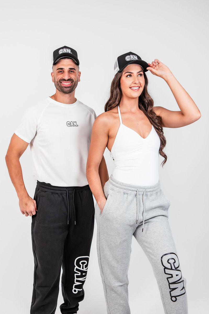 A man and a woman modelling matching white athletic apparel with black and gray accents. The man wears a white short-sleeve t-shirt with a black 'CAN logo on the chest, black sweatpants, and a black CAN. Syndicate Classic Trucker Cap with a white 'CAN' logo.