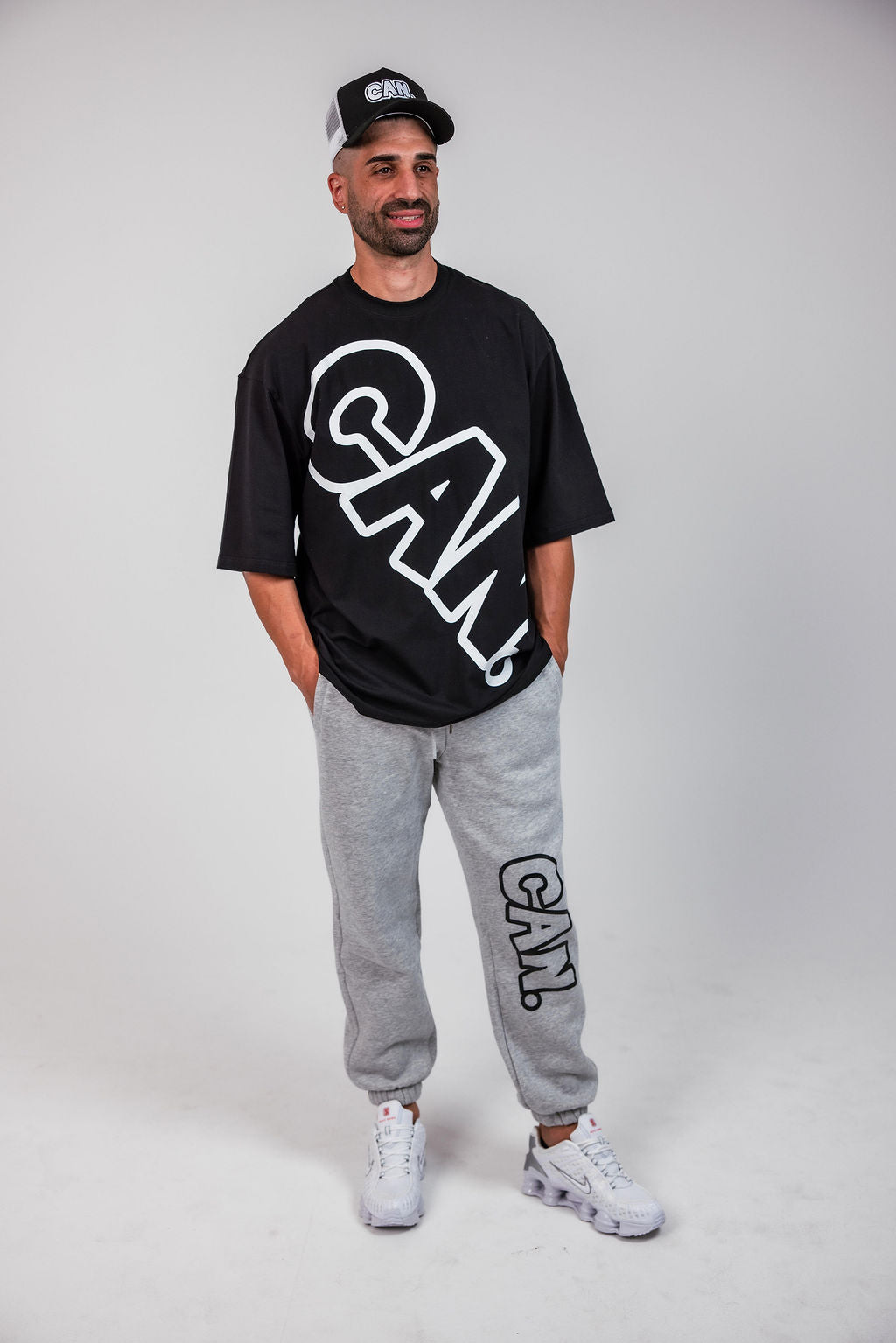 Man wearing black  CAN. Syndicate Bold Oversized Tee with a large logo saying 'CAN" across it as well as a CAN. hat and CAN. Comfort cuffed track pants. 