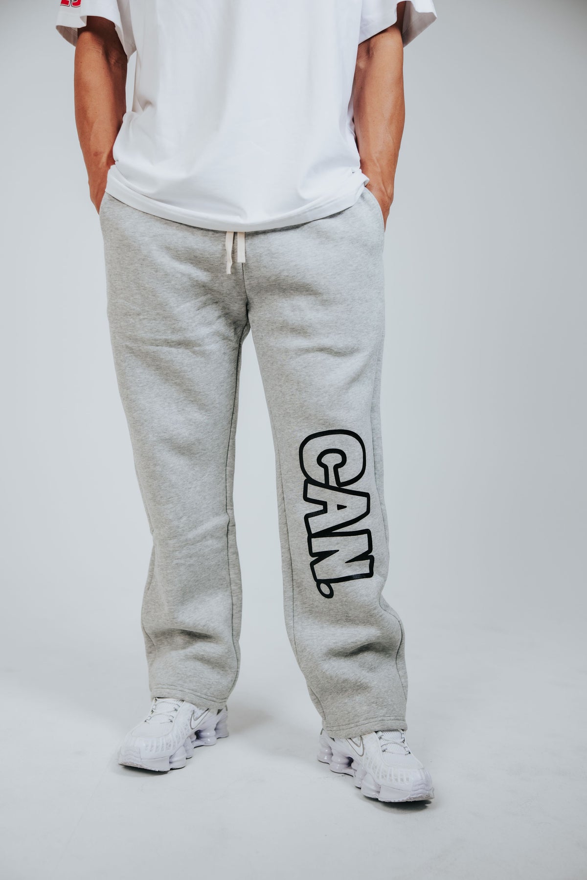 A person wearing light gray CAN. Syndicate Comfort Baggy Track Pants with a large black outlined 'CAN.' logo printed vertically on the left leg, paired with a white t-shirt and white sneakers, shown from the waist down on a light gray studio background.