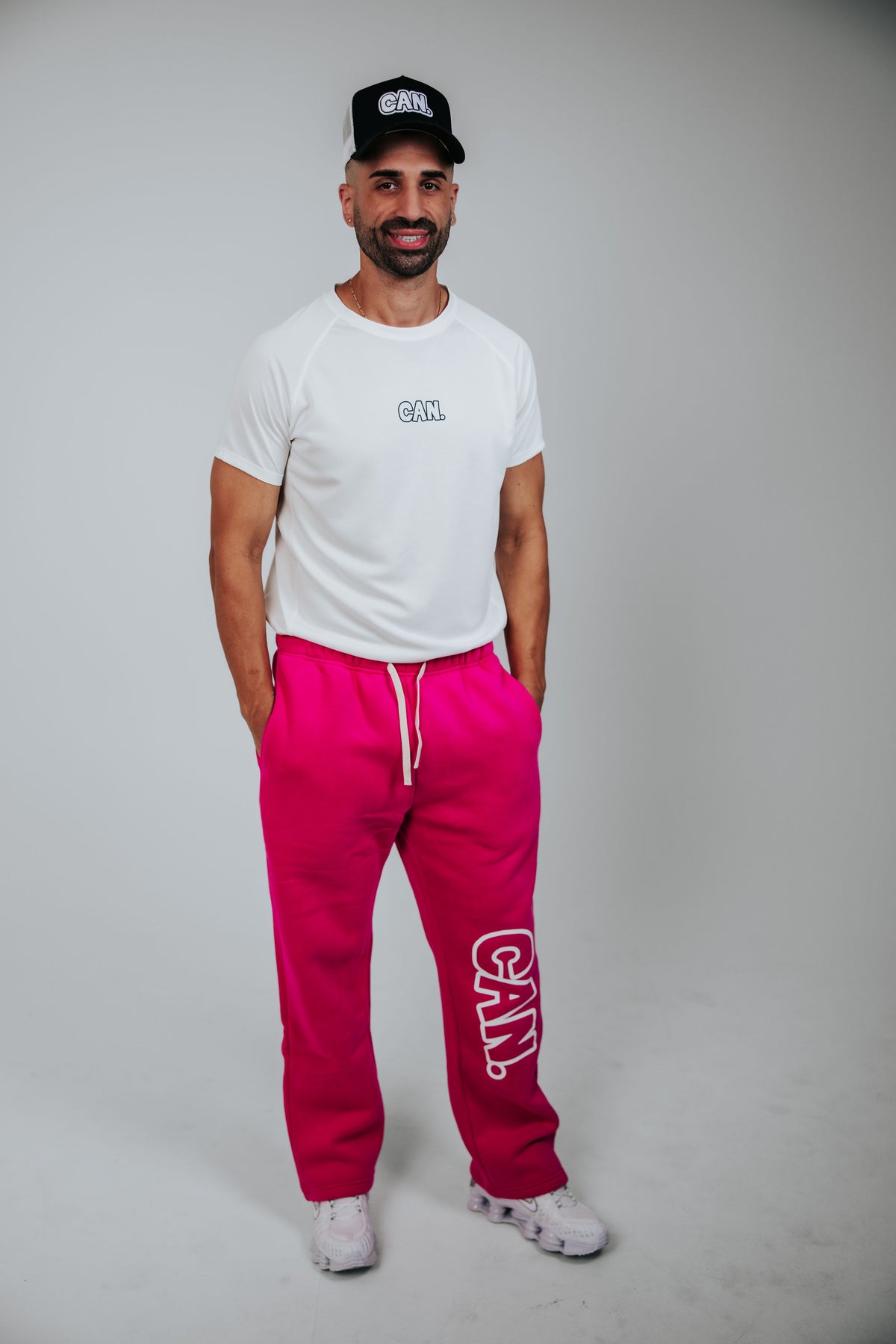 James Webb modelling a white eCAN. Men's Athletic Tee and bright pink CAN. Syndicate Comfort Baggy Track Pants with white drawstrings and a large white 'CAN.' logo printed down the left leg, paired with a black CAN Trucker cap and white sneakers, against a light gray studio background.