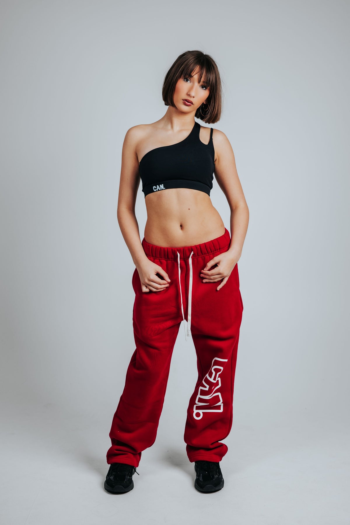 A model stands against a light gray studio background wearing a black CAN. asymmetrical crop top with a small white logo that says CAN. and red CAN. Syndicate Comfort Baggy Track Pants with white drawstrings and a large white vertical logo that says CAN. on the leg.