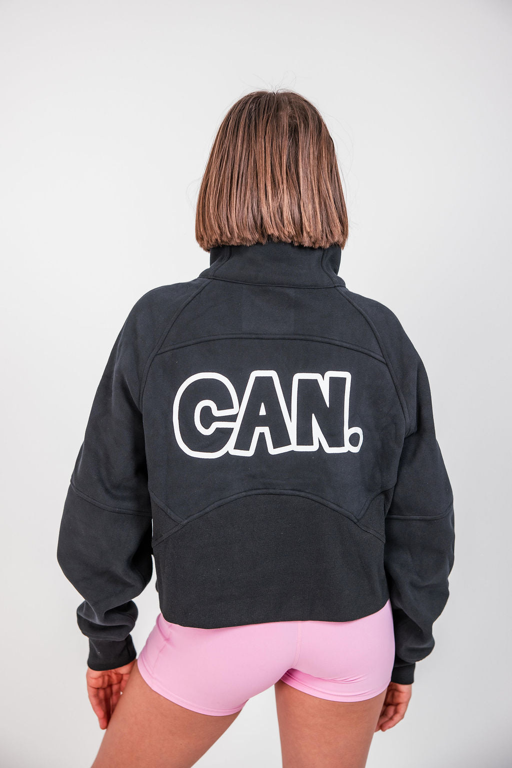 A woman viewed from behind  a Can. Syndicate Cropped Zip Jumper with a white outlined 'CAN.' logo across the back, paired with light pink athletic shorts, against a plain light gray studio background.