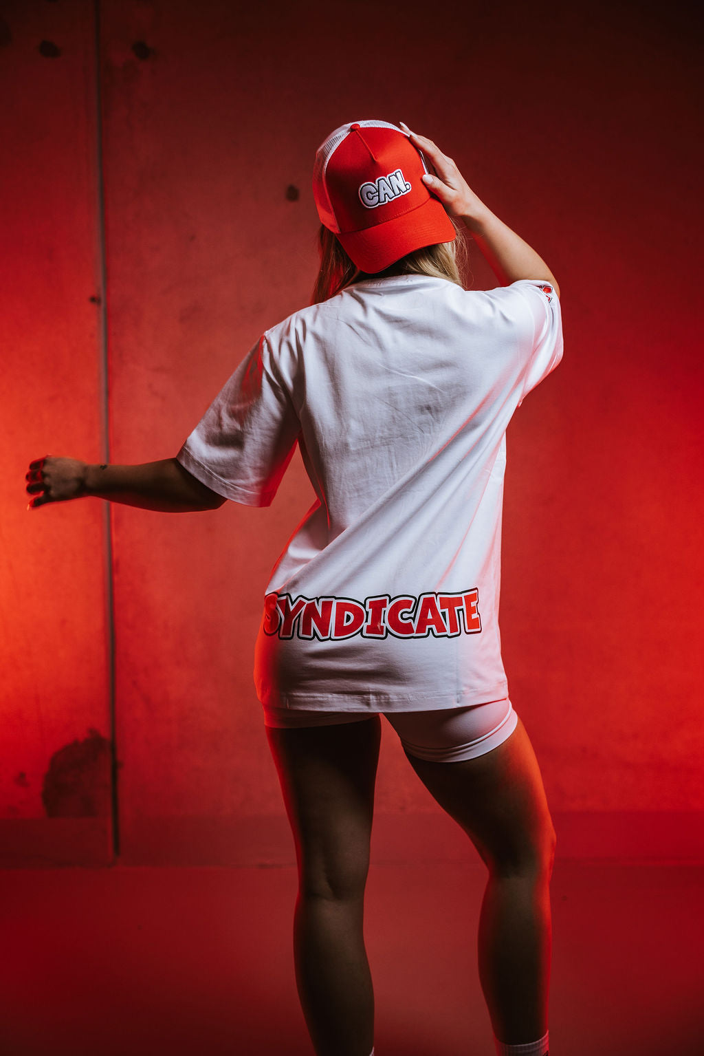 woman facing a red wall wearing a white Can. Syndicate Statement Tee with red "syndicate" logo, a red CAN. Syndicate Classic Trucker Cap and white CAN. Contour Sculpt Shorts. 