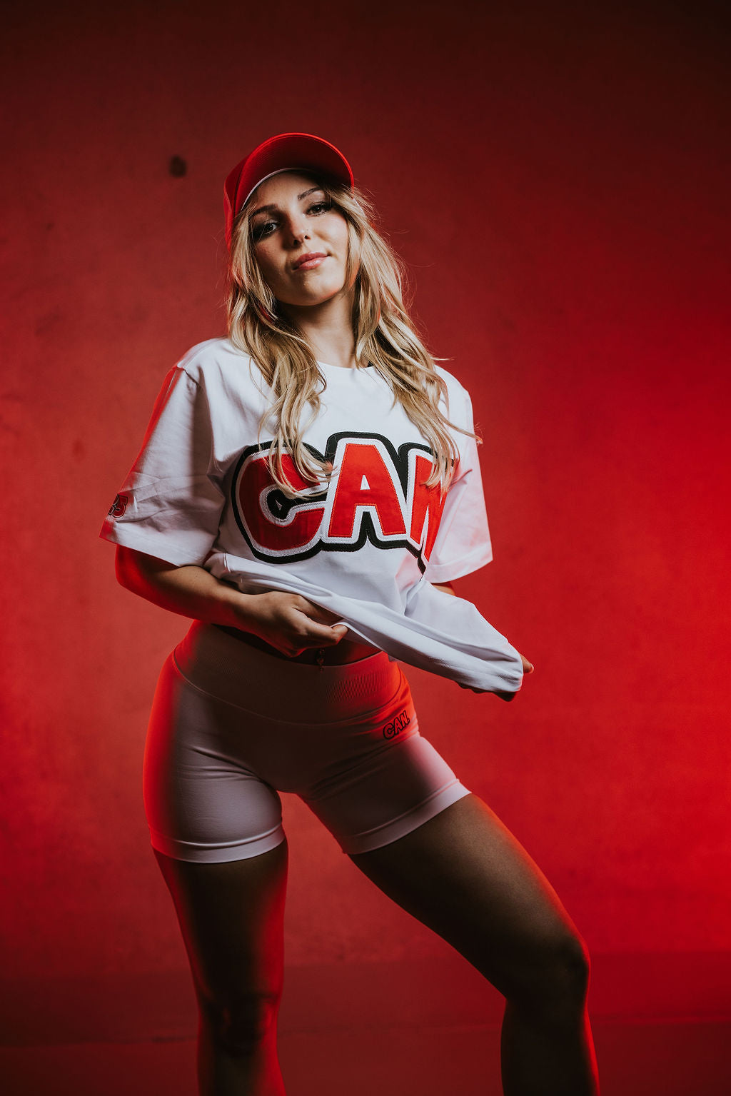 a woman wearing a Can. Syndicate Statement Tee with large red CAN logo, white CAN. Contour Sculpt shorts and a red Can Trucker Cap on a red background. 