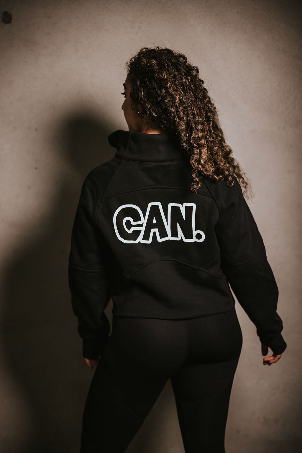 Woman facing a concrete wall wearing a Can. Syndicate Cropped Zip Jumper with a half zip from the brand CAN. She is also wearing Can. black leggings and white crew 