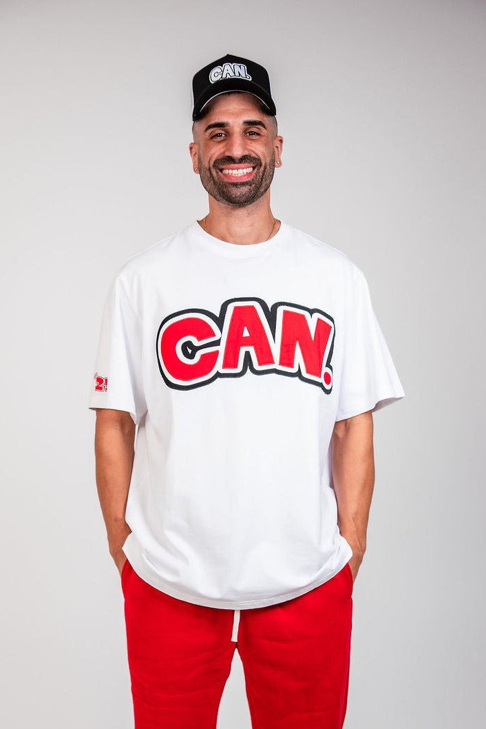A man wearing a black CAN. Syndicate Classic Trucker Cap, white shirt and red track pants. 