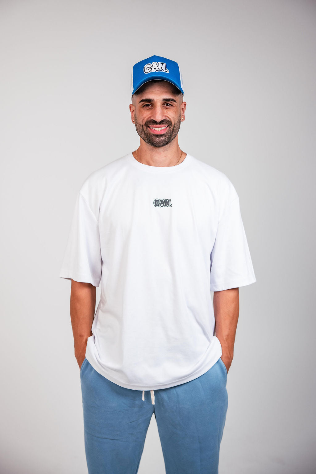 A man standing in front of a white background wearing a blue CAN. Syndicate Classic Trucker Cap with a 'CAN.' logo. he is also wearing a white 'CAN.' shirt and blue 'CAN.' track pants 