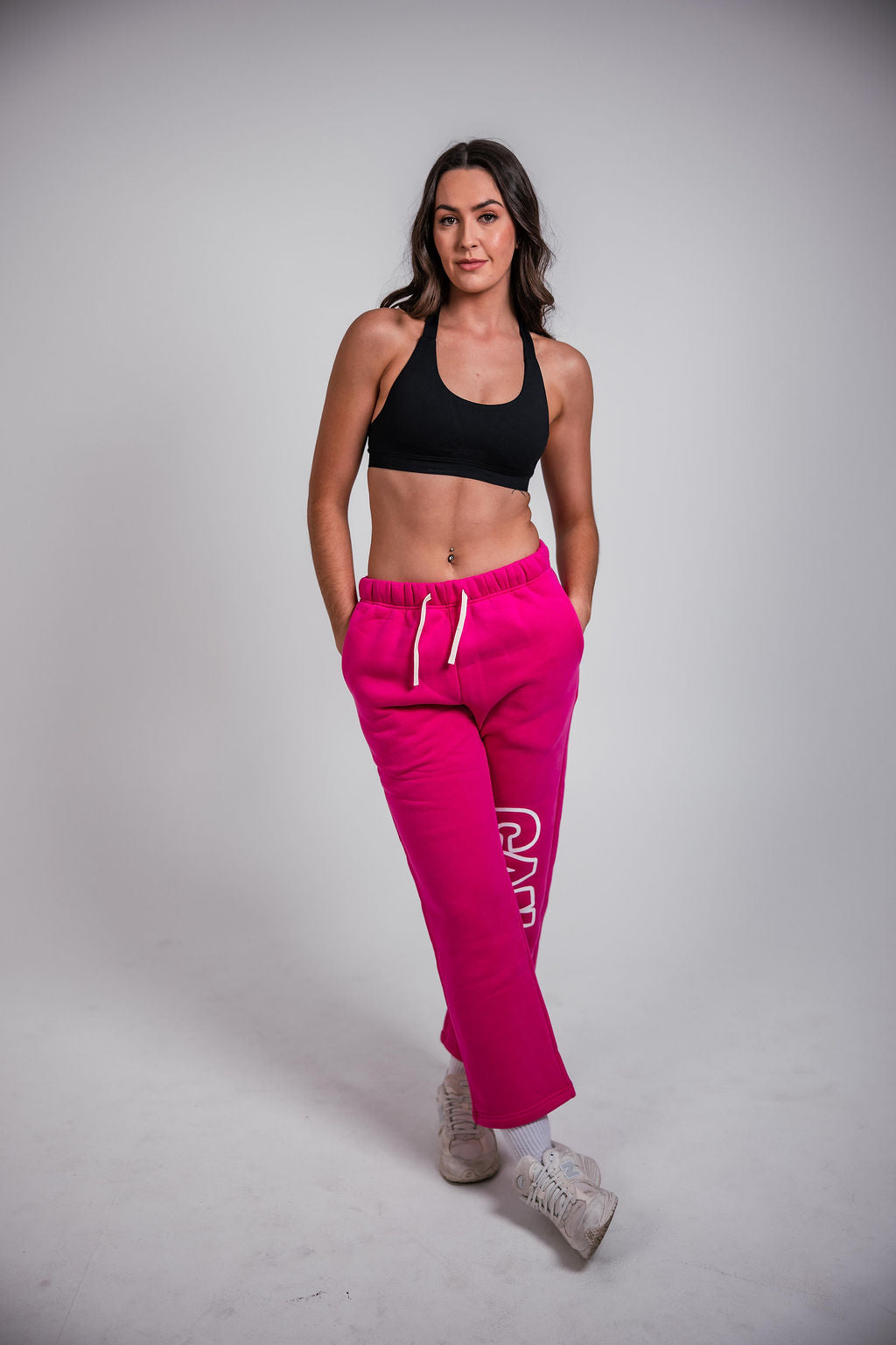 Woman modelling a black CAN. contour sculpt crop top and bright pink CAN. Syndicate Comfort Baggy Track Pants with white drawstrings and a large white 'CAN.' logo printed down the left leg, against a light grey studio background.