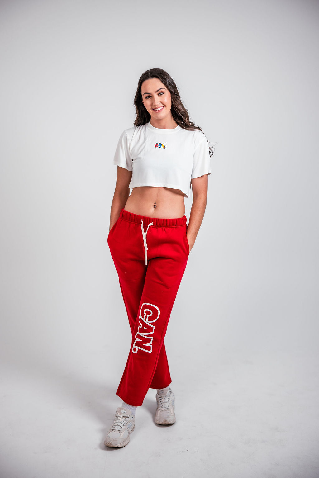 a woman standing with her hands in her pockets wearing a white CAN cropped cover tee and red CAN. Syndicate Comfort Baggy Track Pants with a large 'CAN.' logo on them. 