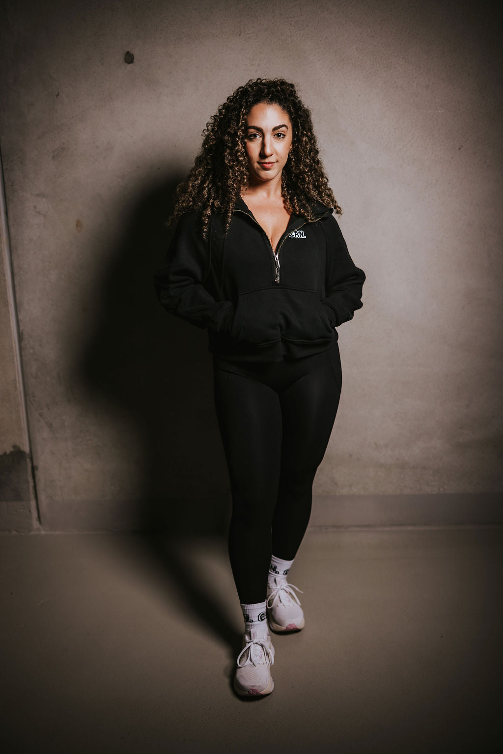 Woman standing in front of concrete wall wearing a Can. Syndicate Cropped Zip Jumper  with a half zip from the brand CAN. She is also wearing Can. black leggings and white crew socks. 