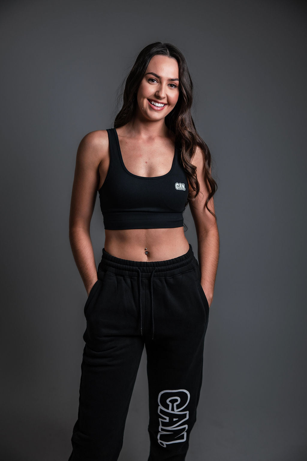 A woman modelling a black Can. Syndicate Forge Crop Top and matching black sweatpants with white 'CAN.' branding, set against a dark grey studio background.