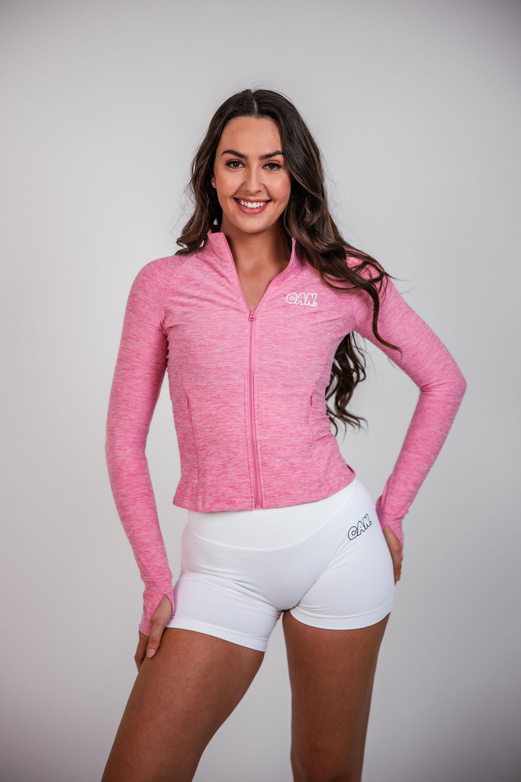 A woman wearing a zipped CAN. Syndicate warm up jacket with white bike shorts with CAN branding in front of a light grey background