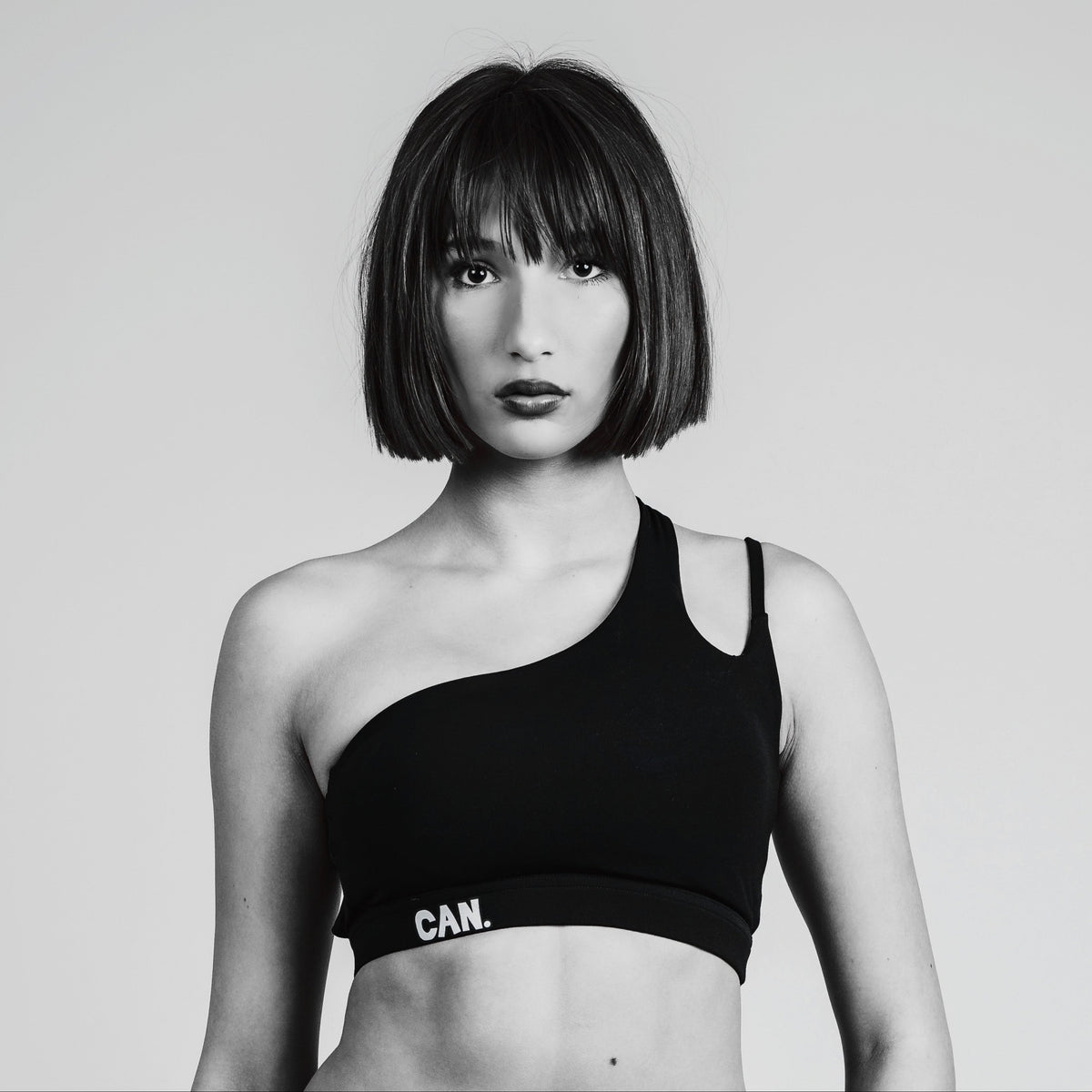 A black one-shoulder sports bra with a wide underband featuring the white text 'CAN.' on the left side, modeled by a woman with a short dark bob haircut against a light gray background.