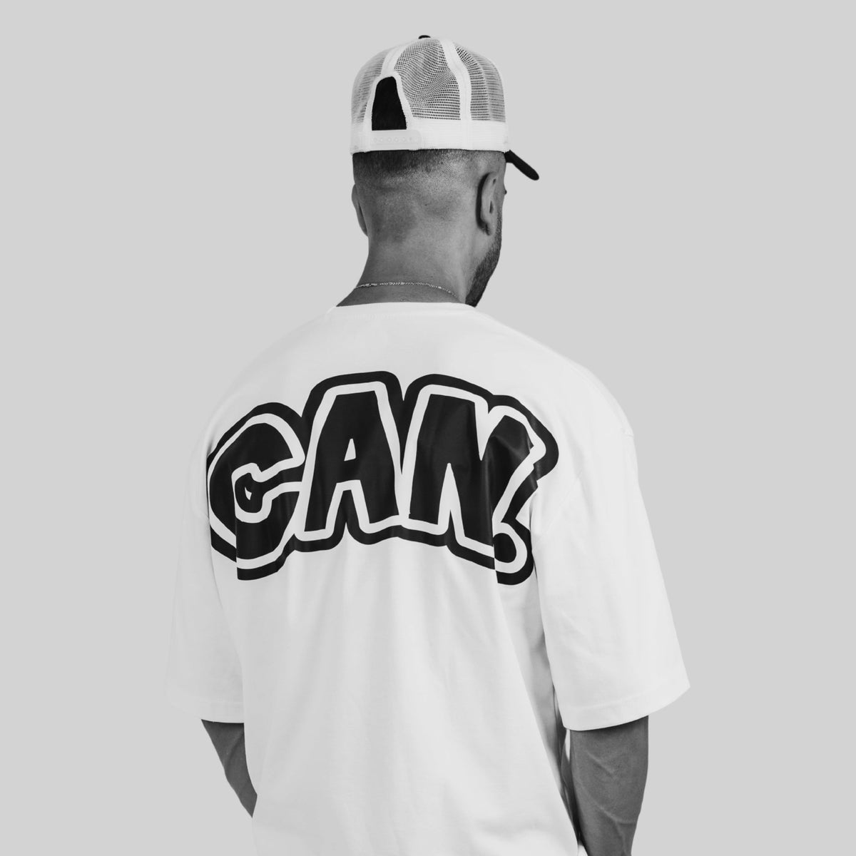 A man viewed from behind wearing a white t-shirt with a large black and white 'CAN' graphic on the back, paired with a gray and white trucker hat, against a solid light gray background.
