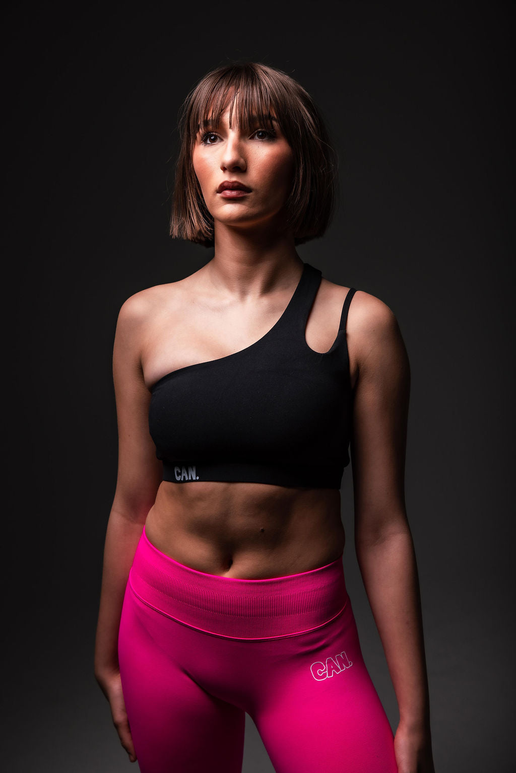 Woman wearing CAN. Syndicate Asymmetrical Cut-Out Crop Top and CAN. Contour Sculpt leggings with a dark grey background. 