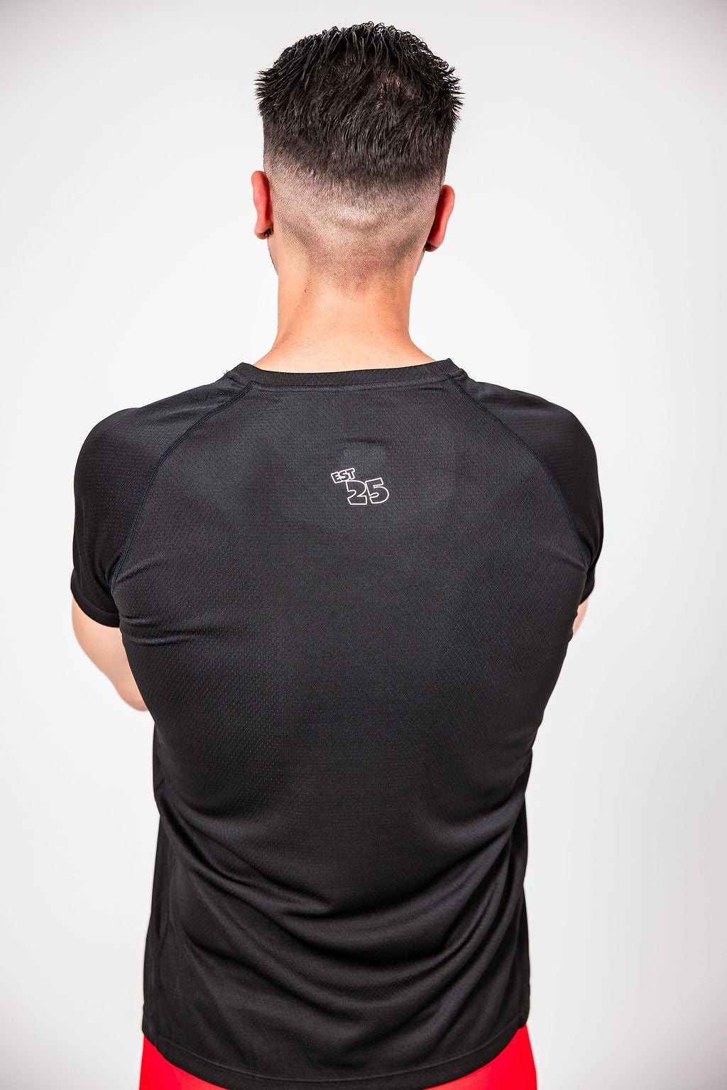 A man facing a white wall wearing a black CAN. Syndicate Men's Athletic Tee  with a small logo that says "est 25" 